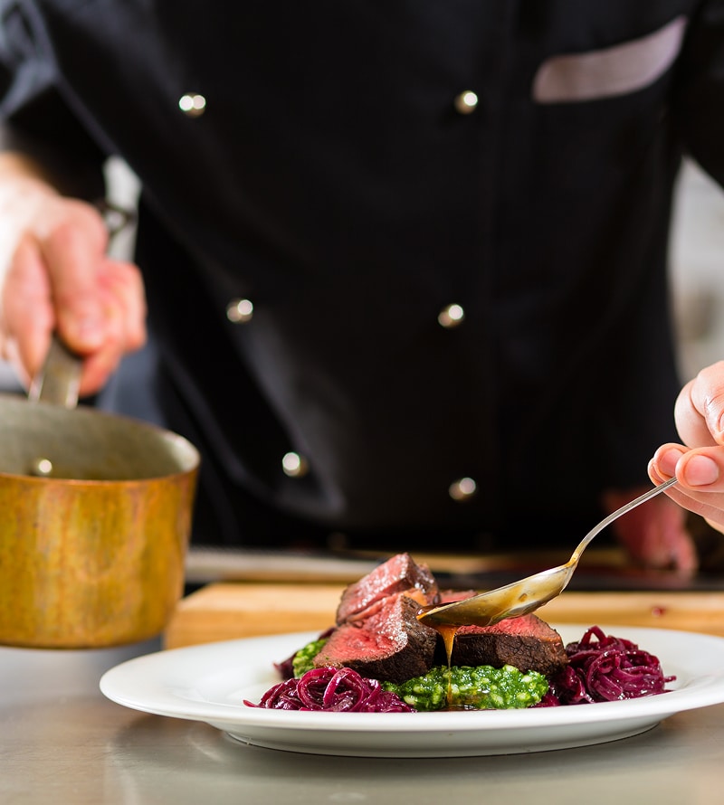 Chef en tablier noir dressant un plat de viande, purée verte et chou rouge, nappant de sauce à la cuillère.