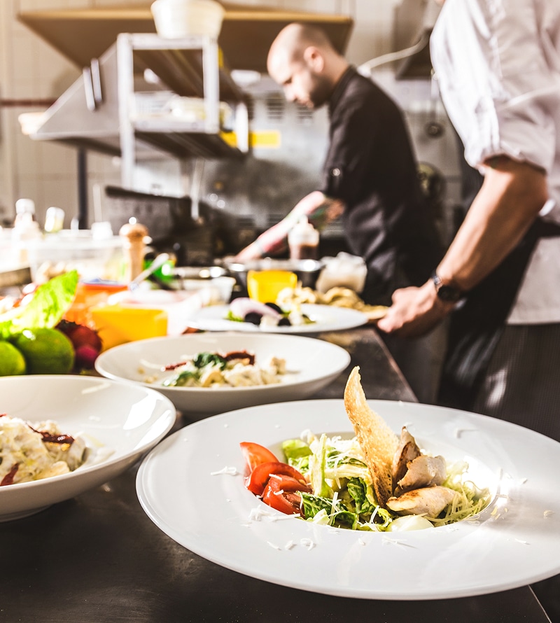 Plats de salades gourmandes prêts sur un comptoir, avec deux chefs affairés en arrière-plan dans une cuisine professionnelle.