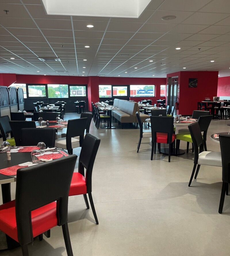 Intérieur moderne de grand restaurant aux murs rouges. Tables dressées, chaises noires/rouges/vertes. Banquettes beiges. Enseigne JYSK visible.
