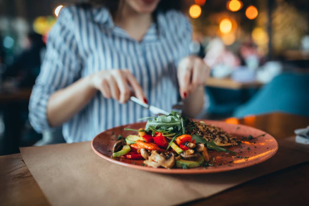 Femme en chemise rayée mangeant un plat végétarien équilibré avec légumes grillés, lentilles et roquette dans un restaurant cosy.