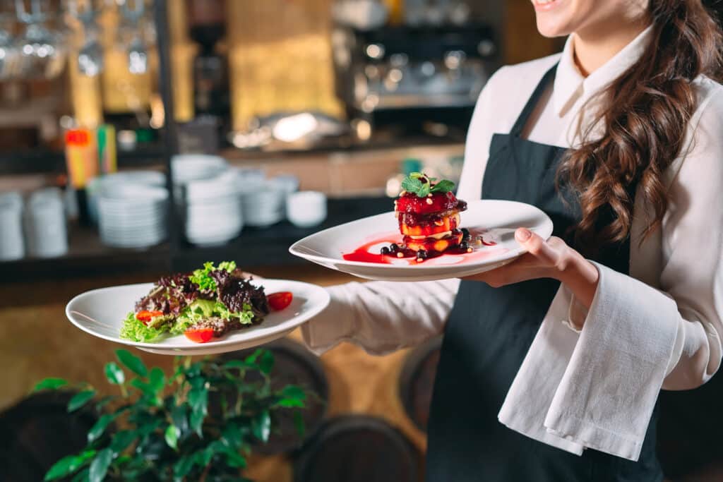 Serveuse souriante en tablier noir, présentant un dessert gastronomique aux fruits rouges et une salade fraîche dans un restaurant.