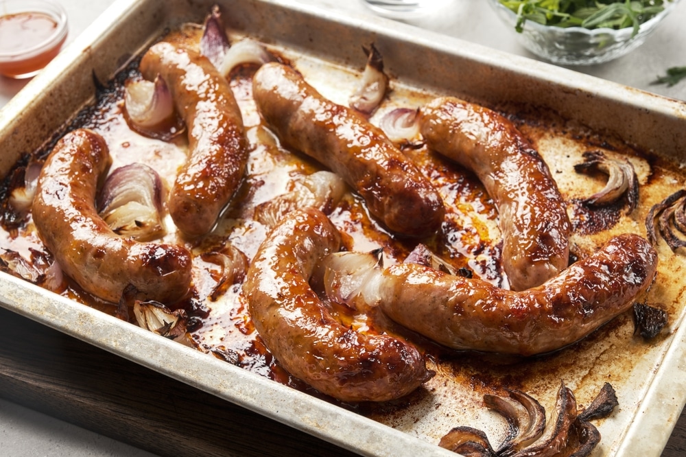 Saucisses Rôties au Four et Oignons Caramélisés Saucisses grillées juteuses et oignons rouges caramélisés sur une plaque de cuisson au four.